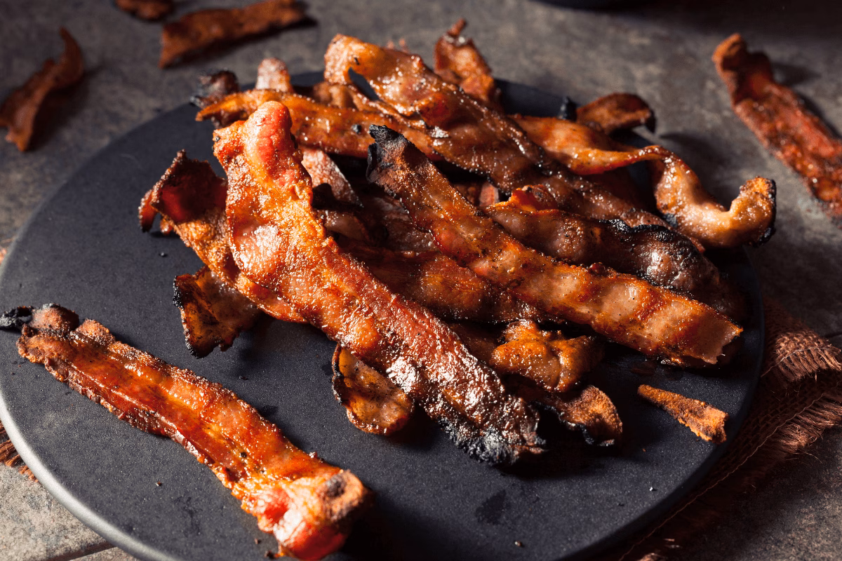 Tiras de bacon frito em um prato escuro, ilustrando gorduras saturadas e inflamatórias que prejudicam a regeneração da pele tatuada.