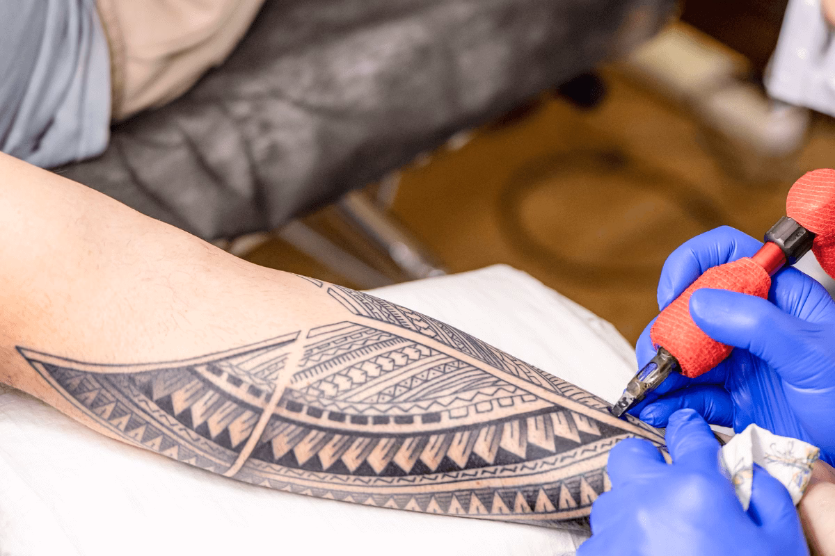 Tatuador aplicando tinta na derme do antebraço com máquina rotativa e luvas azuis.