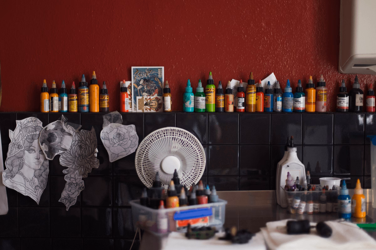 Bancada de estúdio de tatuagem com fileira de tintas coloridas, decalques fixados na parede de azulejo preto e materiais de trabalho organizados.