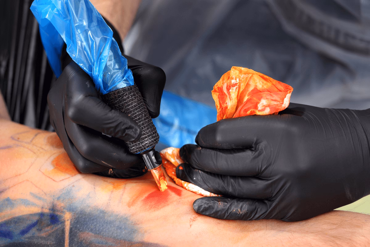 Close-up de tatuador aplicando tinta colorida (laranja e vermelha) na pele, ilustrando o trauma maior que exige uso de sabonete suave e cuidados redobrados na cicatrização de tatuagens coloridas.